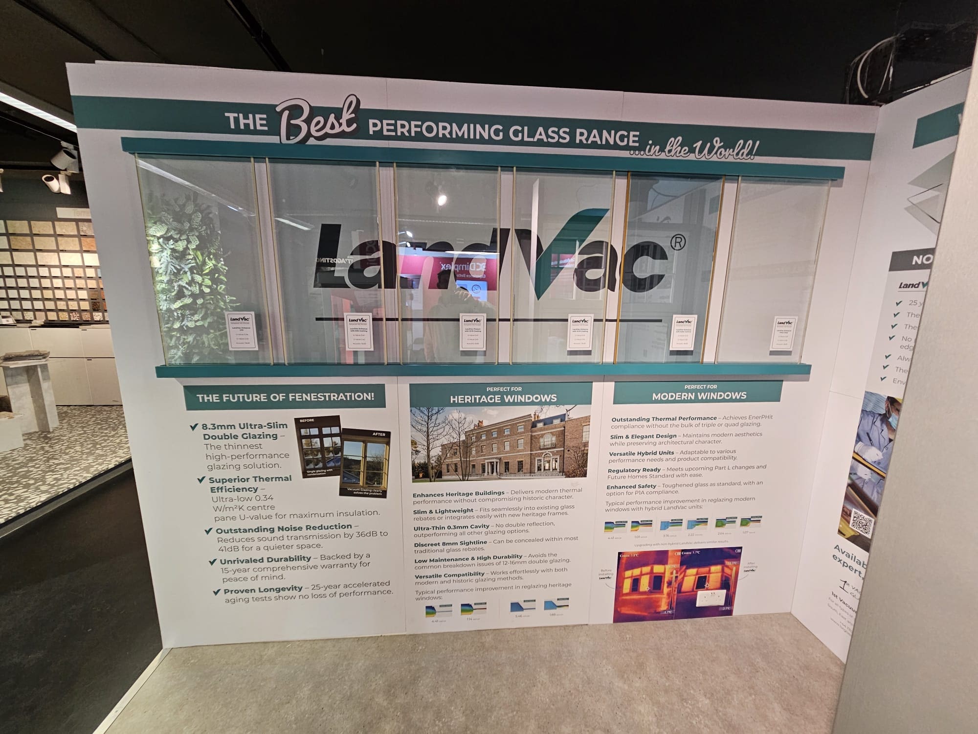 A LandVac exhibition stand at The Building Centre in London, featuring vacuum glazing samples, detailed product information, and promotional graphics on thermal performance and heritage suitability. A Gowercroft Joinery window fitted with LandVac glazing is also on display.