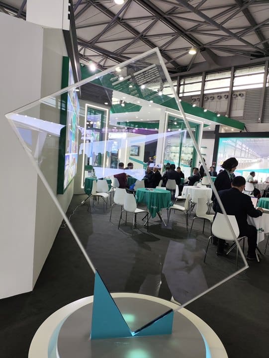 A large diamond-shaped sheet of ultra-clear vacuum glazing on display at an exhibition. The glass is mounted on a round base, allowing attendees to see its transparency and clarity against the backdrop of a modern event space.