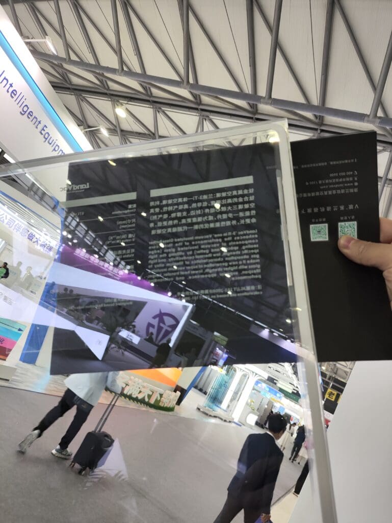 A person holding a transparent LandVac vacuum glass panel at a trade show, demonstrating its clarity and reflectivity. The background shows a modern exhibition hall with display booths, visitors, and signage, highlighting the advanced technology and applications of the glazing product.
