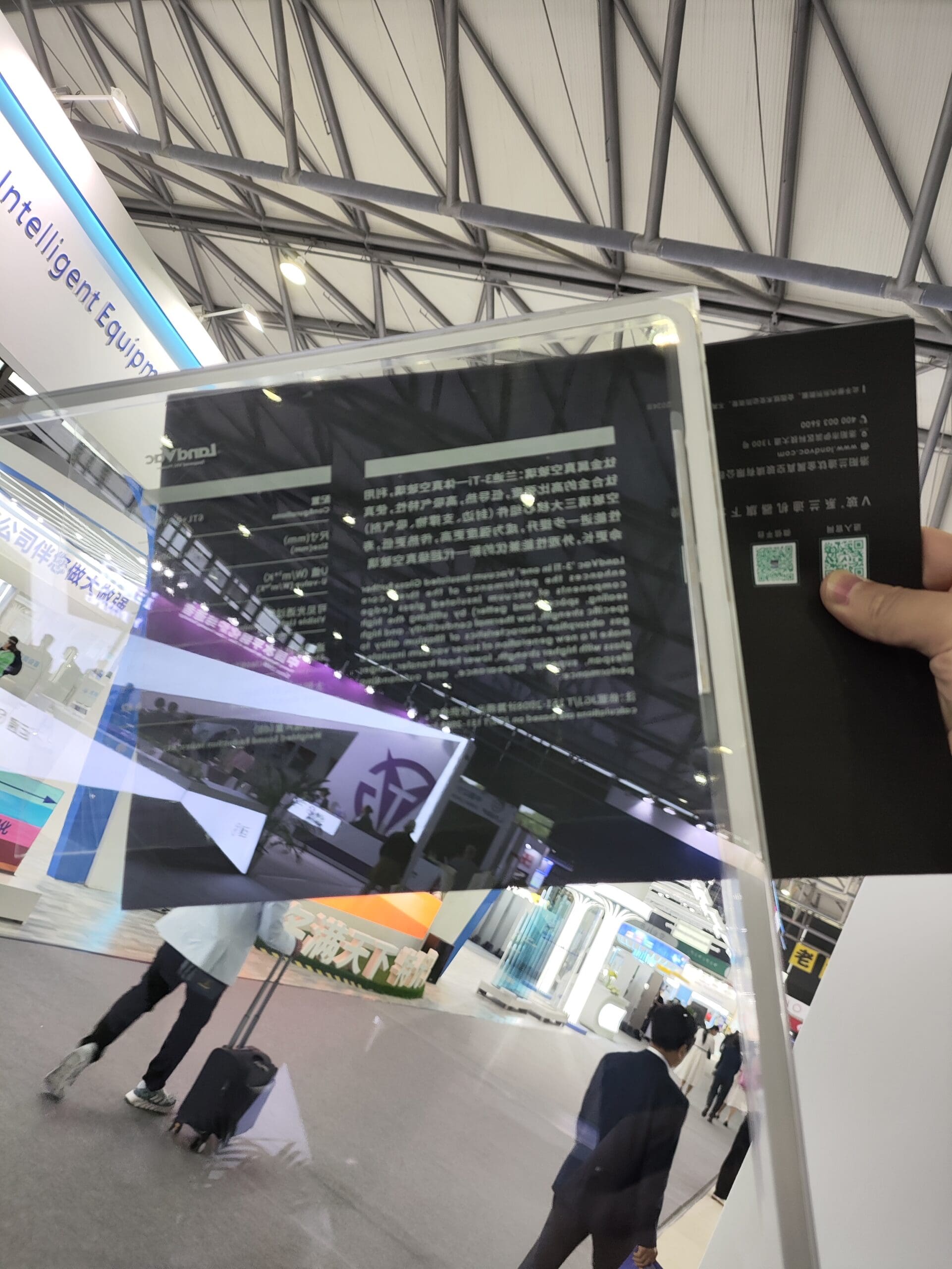 A person holding a transparent LandVac vacuum glass panel at a trade show, demonstrating its clarity and reflectivity. The background shows a modern exhibition hall with display booths, visitors, and signage, highlighting the advanced technology and applications of the glazing product.