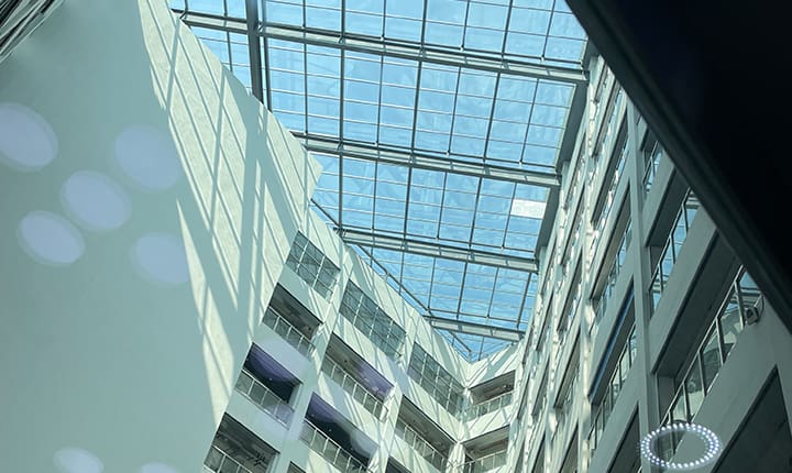 View looking up through a large glass skylight in the central atrium of a modern multi-storey building. The roof is constructed with a grid of clear vacuum-glazed panels, allowing natural light to flood the space. Shadows and reflections are cast onto the white interior walls and balconies below.