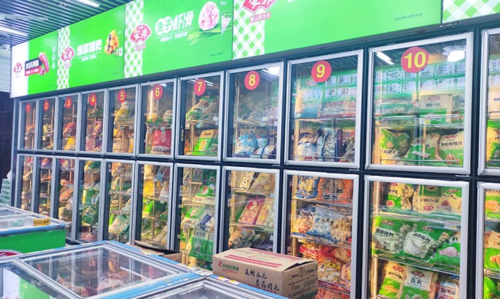 A row of upright commercial freezer units in a brightly lit supermarket, each with clear vacuum-glazed doors displaying frozen food products. Red number labels (1–10) mark each section, and green signage runs above with Chinese branding. A cardboard box sits on the floor in front of the freezers.